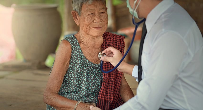 Pflegekraft prüft Puls einer sitzenden älteren Frau.